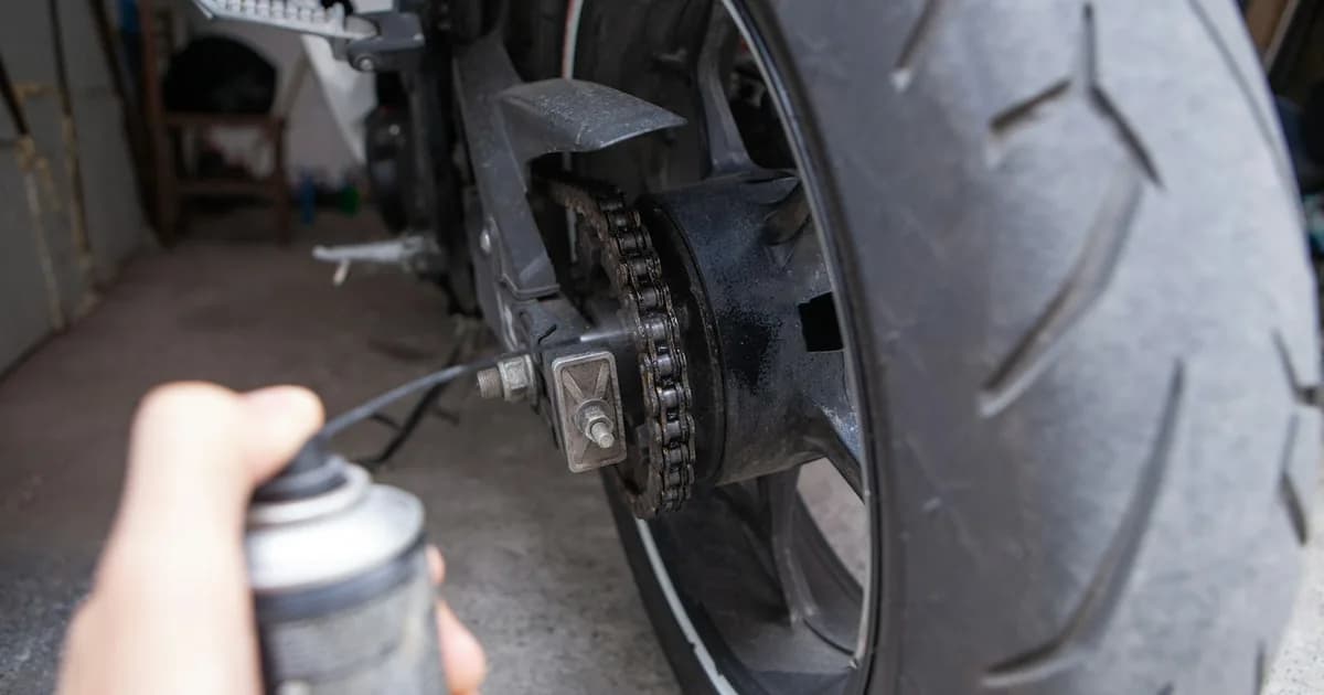 Mão aplica spray de lubrificante na corrente e na coroa de uma moto, com a névoa do jato visível